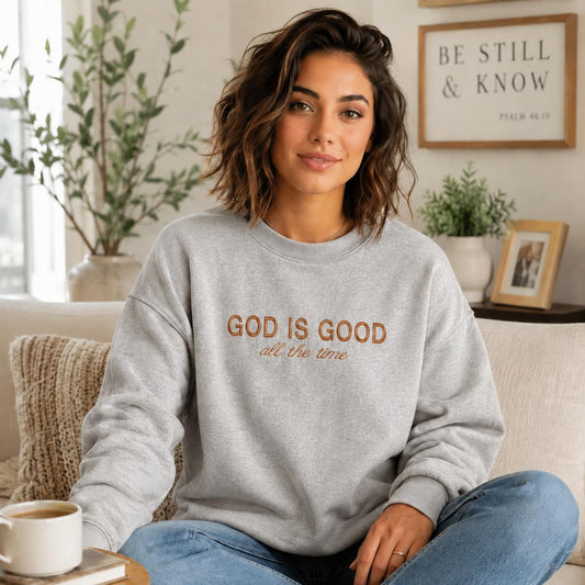 A woman sitting on a beige couch wearing a light gray crewneck sweatshirt with the text 'GOD IS GOOD all the time' in brown lettering. The background shows a cozy living room setting with a framed sign on the wall, a coffee cup, and plants.