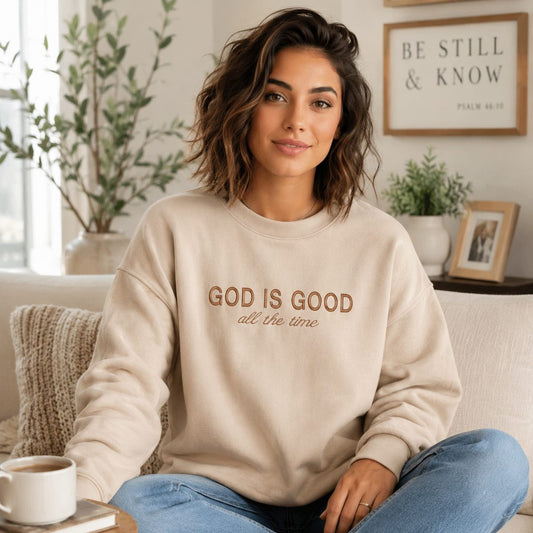 A woman sitting on a couch wearing a beige crewneck sweatshirt with the text 'GOD IS GOOD all the time' in brown lettering. The background shows a cozy living room setting with a coffee cup, plants, and framed wall art.