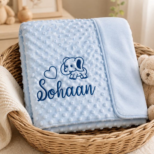 A folded light blue minky dot blanket with a dark blue embroidered elephant, heart, and the name 'Sohaan', displayed in a woven basket next to a teddy bear.
